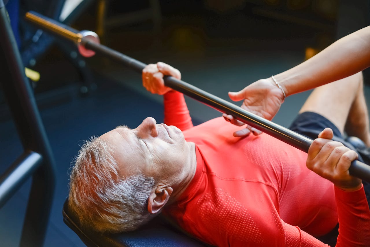 contact-img An elderly man performs a bench press exercise with assistance in a gym, promoting fitness and healthy living.